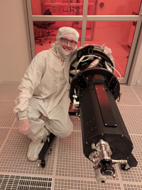 Smiling man in white cleanroom suit crouches beside a large, dark scientific instrument in a red-lit lab.