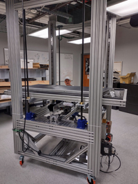 Multi-level silver aluminum experimental frame with black rods, blue components, and a person in a white cap behind.