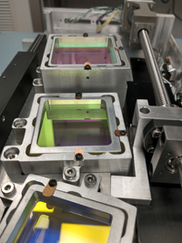 Three rectangular optical modules in metallic frames show vibrant green, purple, and yellow reflections in a lab setup.