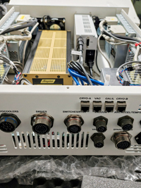 White electronic equipment, top open, showing circuit boards, gold module, blue wires. Labeled multi-pin connectors on front.