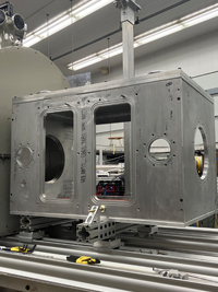 Large silver rectangular metal casing with cutouts, positioned on an assembly track in a manufacturing facility.
